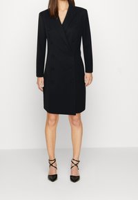 Svart dubbelknäppt kappa med långa ärmar, strukturerad form och textil med struktur. Bärs över åtsittande svarta skor med korsande remmar.