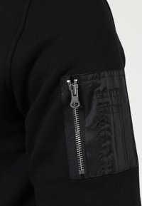 Maglietta nera a maniche lunghe con tasca zip e dettagli di texture del tessuto sulla manica, che accentuano un design a strati.