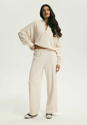 Žena s dlhými tmavými vlasmi, oblečená v béžovej mikine na zips a zodpovedajúcich širokých nohaviciach, stojaca s jednou rukou na bokoch, čierne tenisky.