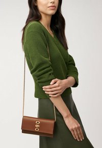 Sac en bandoulière en cuir brun avec une chaîne. Il dispose d'une fermeture à clip avec quincaillerie dorée. Porté avec un pull en tricot vert et une jupe noire.