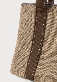 Γωνία από μπεζ και καφέ υφαντό υφασμάτινο τσάντα με σκούρο καφέ λουρί, διαθέτοντας λεπτομέρεια από λευκή ραφή σε απλό φόντο.