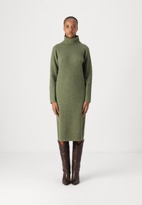 Ελιόχρωμο ριπ πλεκτό φόρεμα με ψηλό γιακά και μακριά μανίκια, φορεμένο με καφέ καουμπόικες μπότες που διαθέτουν λεπτομερή σχέδια ραφής.