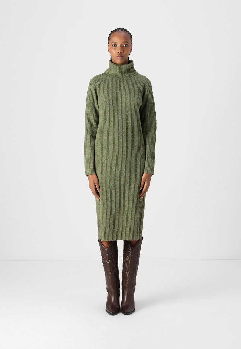 Ελιόχρωμο ριπ πλεκτό φόρεμα με ψηλό γιακά και μακριά μανίκια, φορεμένο με καφέ καουμπόικες μπότες που διαθέτουν λεπτομερή σχέδια ραφής.