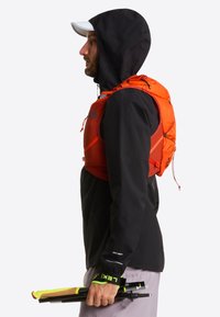 Lekka pomarańczowa kamizelka do nawodnienia z siateczkowymi kieszeniami, na czarnej wodoodpornej kurtce, trzymana przez osobę niosącą kijki trekkingowe.