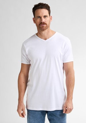 Man met kort krullend haar en baard, die een wit T-shirt met V-hals en een blauwe spijkerbroek draagt, staand tegen een eenvoudige lichte achtergrond.