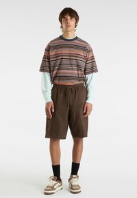 Randig kortärmad skjorta med ficka, tillsammans med bruna shorts med dragsko. Modellen har på sig svarta strumpor och bruna sneakers med vita detaljer.