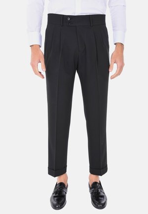 Uomo che indossa pantaloni neri su misura con risvolti, una camicia bianca e mocassini neri con nappine, in piedi davanti a uno sfondo semplice.