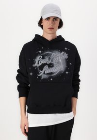Felpa nera con una stampa grafica che presenta "Low Lights Studios" e motivi a stella. Abbinata a una camicia bianca a strati sotto.