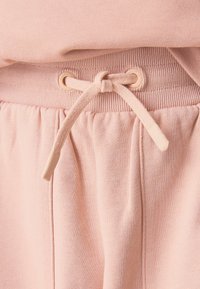 Hele roosad dressipüksid, millel on soonikvöökoht nöörsulguriga ja taskute servades õmmeldud detailid. Pehme kangas.