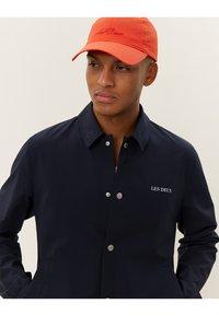 Navy blue outer jacket with snap buttons, featuring the brand name "LES DEUX" in white embroidery. Paired with an orange cap.