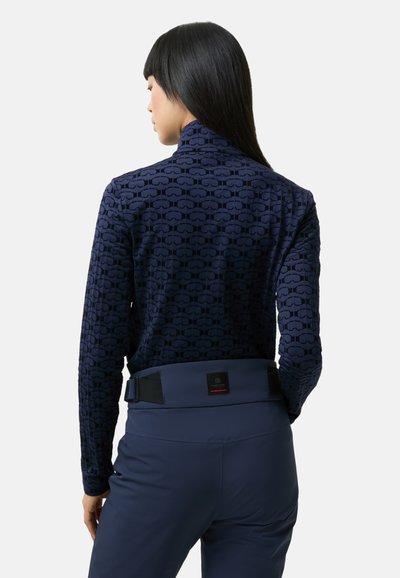 Námořnicky modrý pulovr s texturovaným vzorem brýlí, vysokým límcem a přiléhavým střihem. Kombinován s námořnicky modrými kalhotami s vysokým pasem a s logovou nášivkou.