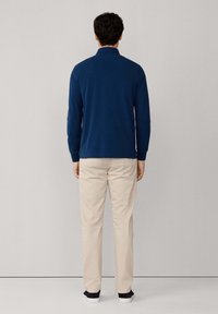 Blauw poloshirt met lange mouwen en een kraag, gecombineerd met beige broek en donkere schoenen, gezien vanaf de achterkant tegen een neutrale achtergrond.