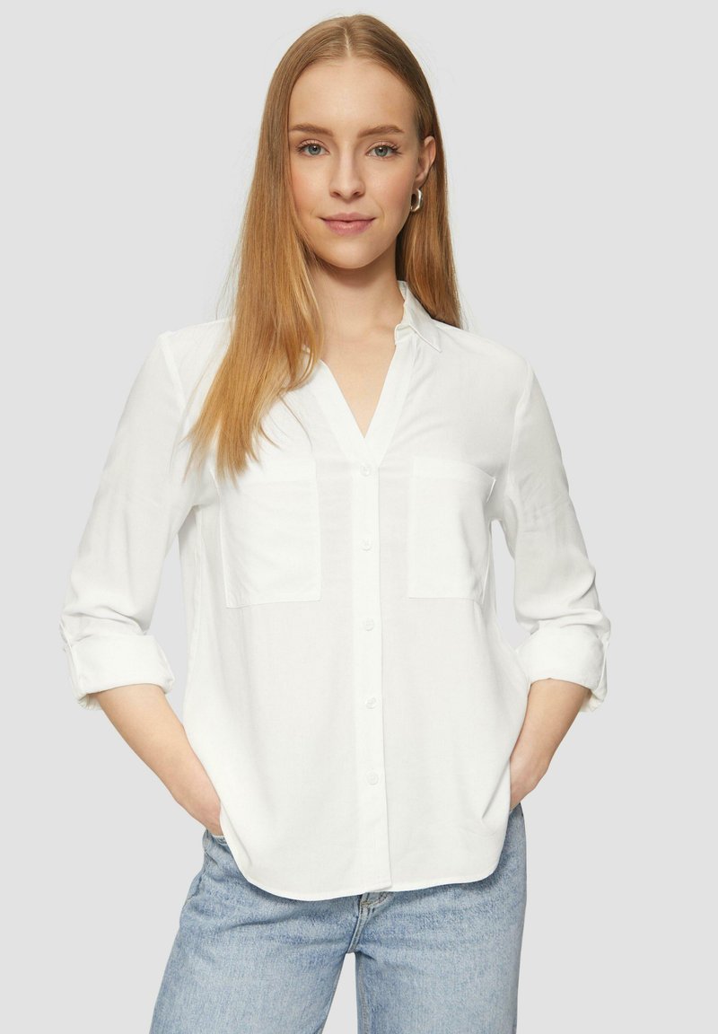 Chemise blanche à col en v, avec deux poches poitrine et des manches retroussées. Tissu léger avec une texture lisse. Associée à un jean bleu.
