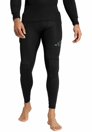Homme portant des leggings de compression rembourrés noirs et un haut à manches longues, debout pieds nus sur un fond blanc.