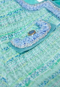 Tessuto tweed verde chiaro e blu con una superficie testurizzata, caratterizzato da una tasca decorativa con bordi arricciati e un bottone con accento dorato.