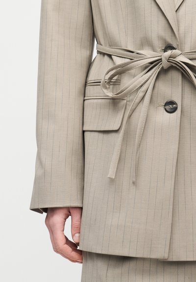 Κοντινό πλάνο μπεζ σακακιού με ρίγες, τσέπη με καπάκι, μαύρα κουμπιά και δεμένη υφασμάτινη ζώνη στ η μέση, με ένα χαλαρό χέρι.