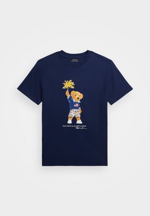 Marineblaues T-Shirt mit einer Grafik eines Bären, der ein Hemd mit amerikanischer Flagge trägt und gelbe Blumen hält. Polo Bear von Ralph Lauren Text darunter.