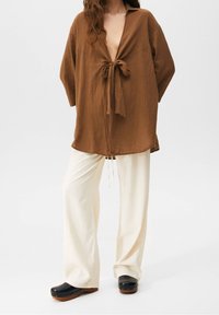 Femme portant un haut long à manches longues marron et ample noué au niveau de la poitrine, un pantalon large crème, et des sabots noirs à semelle en bois, se tenant contre un fond blanc.