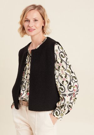 Zwarte textuurvest zonder sluitingen, gedragen over een gestreepte blouse met bloem- en paisleydessins. Model heeft handen in de zakken.
