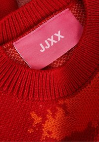 Red knitted sweater with a ribbed collar and vibrant color gradient, featuring a pink label displaying “JJXX” in bold white letters.