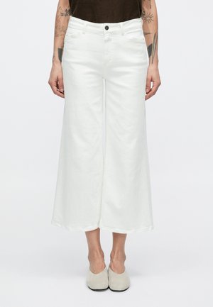 Pantalon blanc à jambe large en coton avec une finition texturée, doté d'une boutonnière frontale et de détails de poches. L'ourlet tombe à mi-mollet.