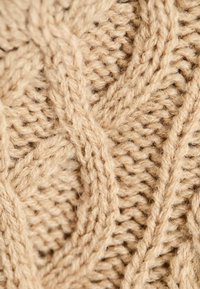 Beige színű kötött textil, texturált kábelmintával, amely vastag fonalakból áll, létrehozva egy réteges, dimenzionális hatást.