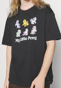 T-shirt noir avec des impressions de personnages colorés de "My Little Pony" dans diverses poses et un logo en texte blanc en bas. Manches courtes, coupe décontractée.