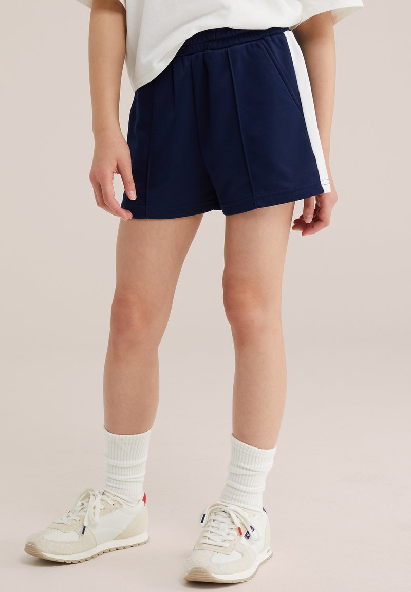 Personne portant un short bleu marine avec des panneaux latéraux blancs, des chaussettes blanches et des baskets blanches, debout sur un fond neutre.
