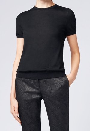 Osoba w czarnej dopasowanej koszulce z krótkim rękawem i czarnych spodniach o fakturze w kwiatowy wzór, z jedną ręką w kieszeni.