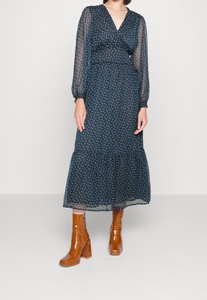 Frau trägt ein marineblaues Midikleid mit langen Ärmeln, kleinen weißen Mustern und braunen hochhackigen Stiefeletten, stehend.