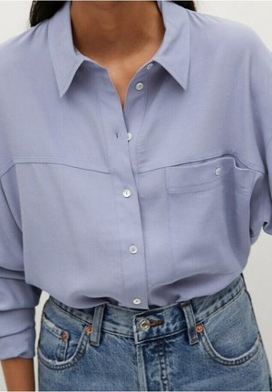 Frau trägt ein hellviolettes Hemd mit Brusttasche, das in hochtaillierte blaue Jeans gesteckt ist.