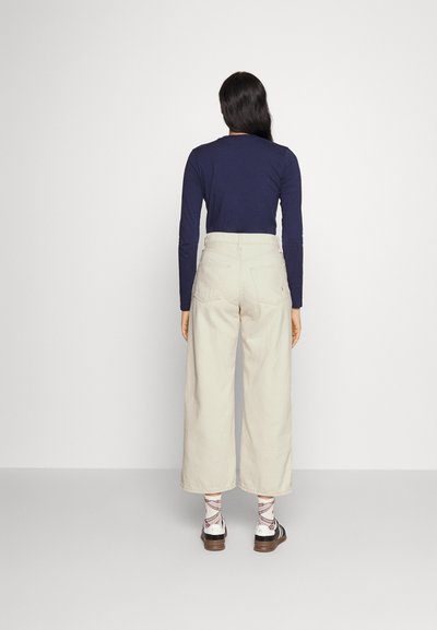 Granatowa koszulka z długim rękawem w połączeniu z kremowymi spodniami z szerokimi nogawkami. Spodnie mają tylne kieszenie i wysoki stan. Uzupełnione wzorzystymi skarpetkami i butami.