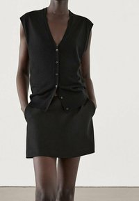 Schwarze ärmellose Strickjacke mit tiefem V-Ausschnitt, Knopfverschluss und passendem schwarzen Rock mit Taschen, beide aus glattem Stoff.