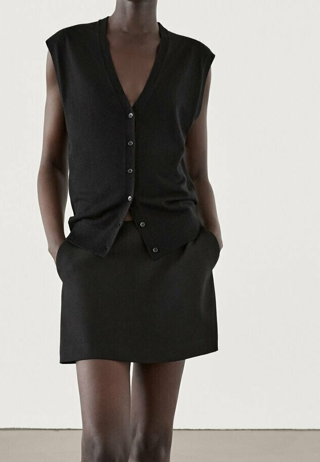 Schwarze ärmellose Strickjacke mit tiefem V-Ausschnitt, Knopfverschluss und passendem schwarzen Rock mit Taschen, beide aus glattem Stoff.