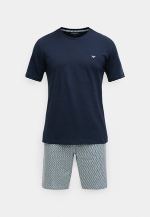 Temno modra bombažna majica s kratkimi rokavi in subtilnim logotipom; kombinirana z lahkimi kratkimi hlačami s geometrijskim vzorcem v beli in modri barvi.