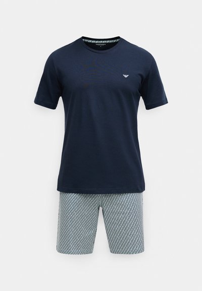 Modré bavlnené tričko s krátkym rukávom a jemným logom; doplnené vzorovanými svetlými šortkami s geometrickým dizajnom v bielej a modrej farbe.