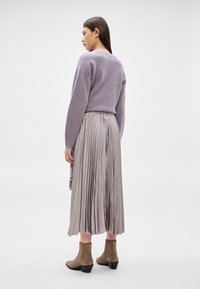 Moteris dėvi levandų spalvos megztą megztinį, sidabro spalvos raukšlėtą midi sijoną ir taupe spalvos kulkšnies aulinukus, stovi neutralaus balto studijos fono priekyje.