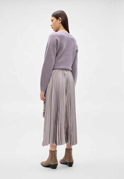 Femme portant un pull en tricot lavande, une jupe midi plissée argentée et des bottines taupe, debout sur un fond studio blanc neutre.