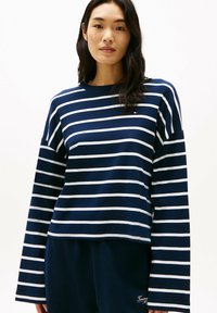 Maglia a maniche lunghe blu navy con strisce orizzontali bianche, dotata di collo a giro e un piccolo logo. Tessuto di cotone morbido.