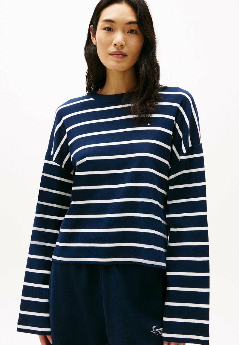 Maglia a maniche lunghe blu navy con strisce orizzontali bianche, dotata di collo a giro e un piccolo logo. Tessuto di cotone morbido.