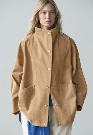 Frau trägt übergroße hellbraune Wildlederjacke mit silbernen Druckknöpfen, einen blauen Strickschal und cremefarbene Hosen vor schlichtem grauem Hintergrund.