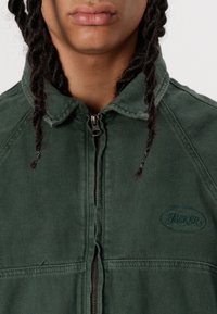 Grøn, lynlåsjakke lavet af slidstærkt stof med en tekstureret overflade, der har et logo-patch og en nedfoldelig krave.