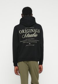 Άτομ ο με την πλάτη στραμμένη φοράει μαύρο φούτερ με κουκούλα, με μεγάλο κείμενο "Originals Studio" και μικρότερη περιγραφική εκτύπωση στην πλάτη.