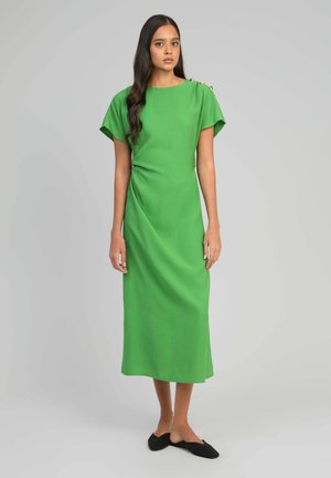 Femme aux longs cheveux foncés portant une robe midi verte avec des détails de boutons sur l'épaule et des chaussures noires sans lacets, se tenant devant un fond gris.