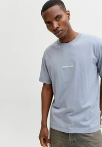 Heleheles sinine puuvillane T-särk lühikeste varrukatega ja ümmarguse kaelusega. Esiküljel on valge käsitsi kirjutatud tekst, sile tekstuur ja mugav lõige.