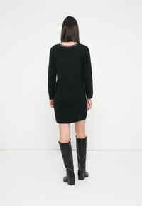 Frau, die ein schwarzes langärmliges Kleid mit Spitzendetails am Ausschnitt und kniehohen schwarzen Stiefeln mit Absatz trägt, steht vor einem weißen Hintergrund.