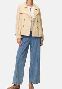 Beige dobbeltbrystet jakke med krave og dekorative knapper, parret med brede blå denimbukser. Letvægtsstof og afslappet pasform.