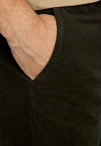 Zwarte corduroypantalon met een gestructureerd oppervlak. De hand is in een zijzak geplaatst, wat de details van de stof en de stiksels toont.