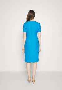 Přiléhavé šaty z jasně modré látky, s krátkými rukávy, délkou sahající ke kolenům, se skrytým zipem na zádech. Hladká textura, minimalistický design.