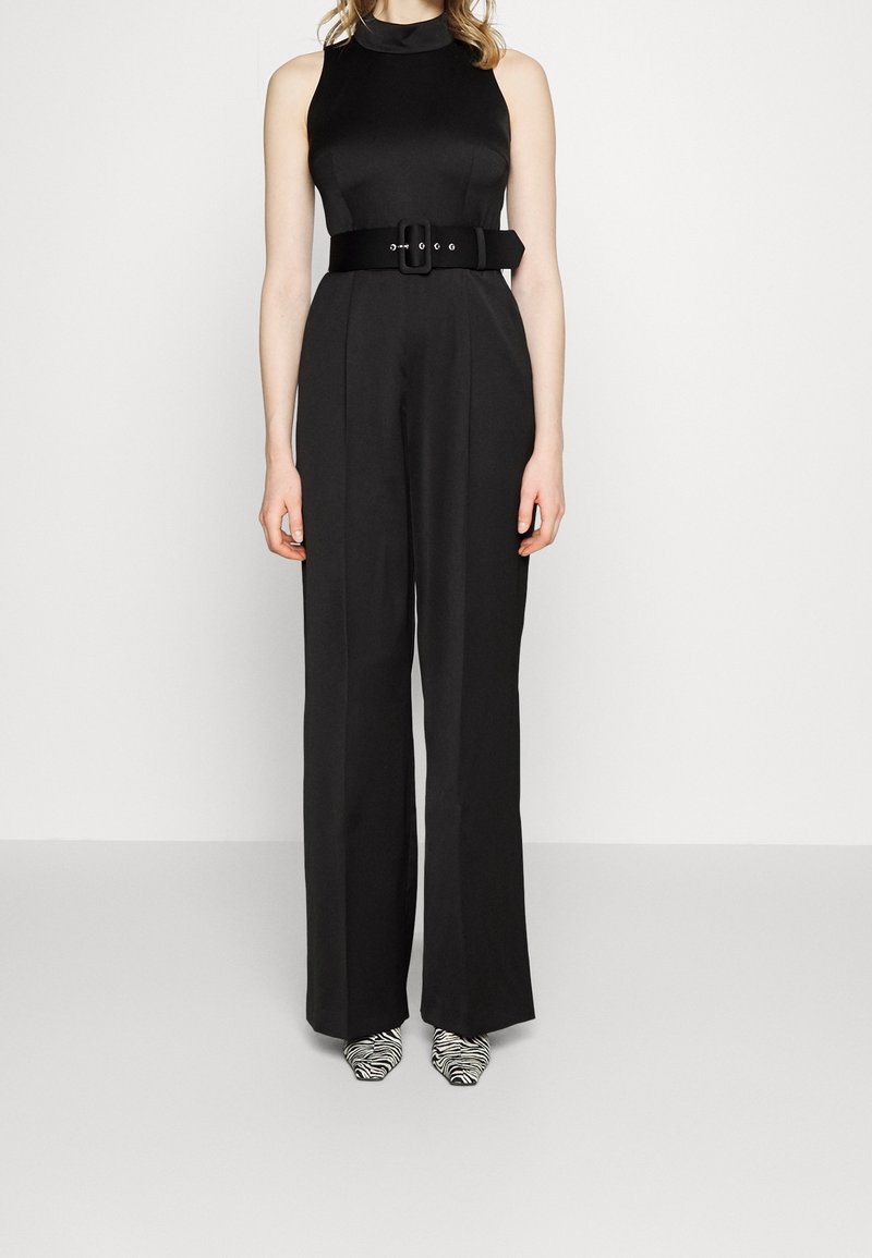 Schwarzer ärmelloser Jumpsuit mit tailliertem Oberteil, weit geschnittenen Hosenbeinen und einem schwarzen Gürtel mit rechteckiger Schnalle. Zebra-print Schuhe vervollständigen das Outfit.